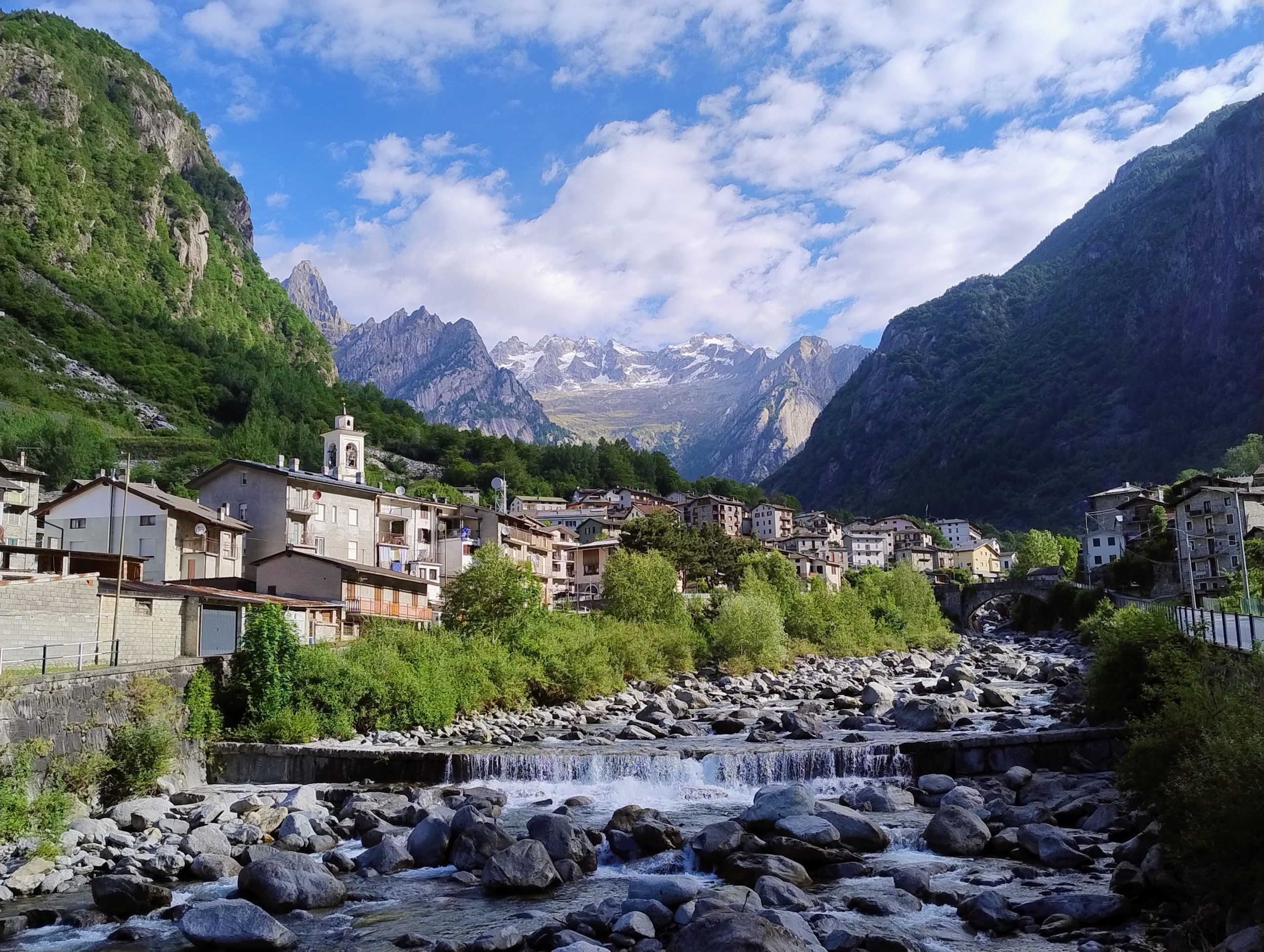 Veduta panoramica di un pittoresco villaggio alpino di montagna incastonato tra alte vette rocciose, con un fiume a cascata e vegetazione lussureggiante in primo piano sotto un cielo azzurro.